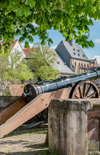Fügeler-Kanone (Ruben Emme) Historische Kanone unter Bäumen mit Blick auf Fachwerkhäuser und eine alte Stadtmauer im Hintergrund.