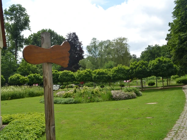 Holzpfad-Schild vor grüner Gartenanlage, gesäumt von Bäumen und Blumenbeeten, Spazierweg an der Seite.