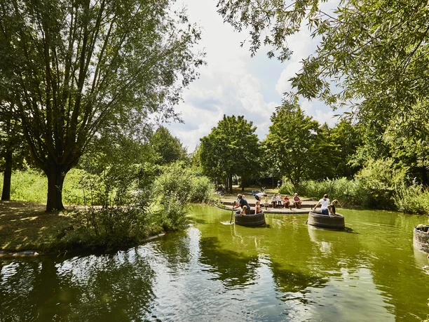 Menschen fahren mit Rundbooten auf grünlichem Wasser durch einen idyllischen, von Bäumen gesäumten Kanal.