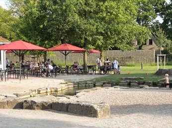 Gastro_Spielerei.jpg Menschen sitzen entspannt in einem Café im Freien mit roten Sonnenschirmen, Bäumen im Hintergrund.