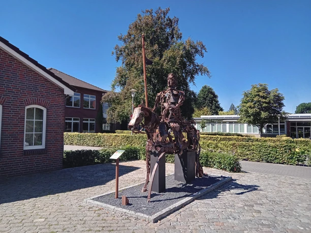 Lieblingsort Rathausmarkt Apen mit Ritter Herbord Eine imposante Metallskulptur eines Ritters auf einem Metallpferd steht auf gepflastertem Platz vor Büschen und Gebäuden, umgeben von klarem Himmel an einem sonnigen Tag.