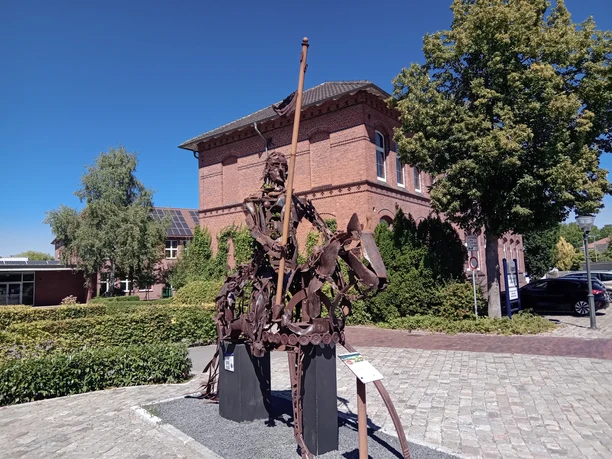 Aper Lieblingsort mit Ritter Herbord Das Bild zeigt eine eindrucksvolle Metallskulptur eines Ritters mit Speer auf einer gepflasterten Fläche vor einem historischen Backsteingebäude. Die Skulptur aus Metallteilen zieht die Blicke auf sich und verleiht dem Platz eine besondere Atmosphäre. Strahlend blauer Himmel und grüne Bäume rahmen die Szene malerisch ein.