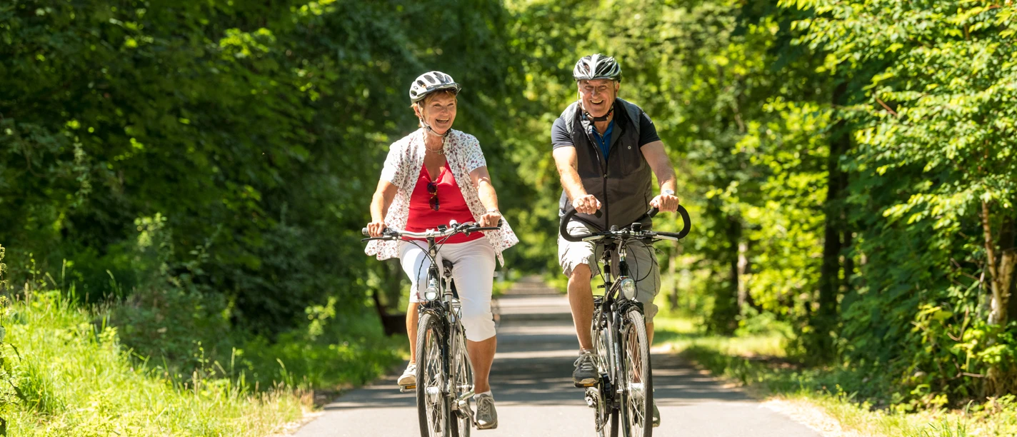 Radfahrer im Bergischen Ein älteres Paar fährt mit Fahrrädern lächelnd auf einem von Bäumen gesäumten Radweg.