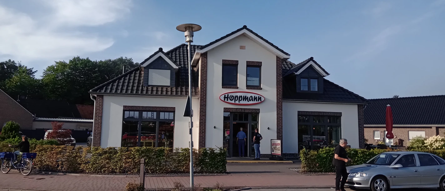 Bäckerei Hoppmann in Augustfehn Das Bild zeigt die Fassade der Bäckerei Hoppmann in Augustfehn. Das Gebäude hat eine traditionelle, einladende Architektur mit einem markanten Schriftzug „Hoppmann“ über dem Eingang. Vor der Bäckerei sind mehrere Personen und geparkte Fahrräder zu sehen.