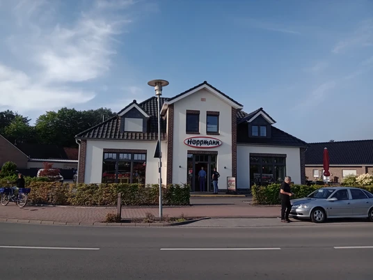 Bäckerei Hoppmann in Augustfehn Das Bild zeigt die Fassade der Bäckerei Hoppmann in Augustfehn. Das Gebäude hat eine traditionelle, einladende Architektur mit einem markanten Schriftzug „Hoppmann“ über dem Eingang. Vor der Bäckerei sind mehrere Personen und geparkte Fahrräder zu sehen.
