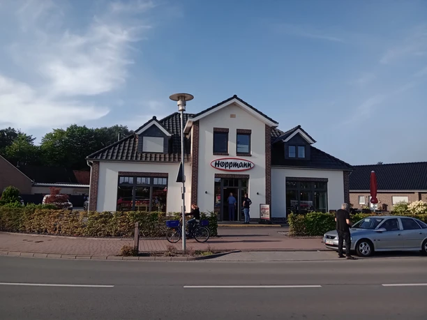 Bäckerei Hoppmann Das Bild zeigt die Fassade der Bäckerei Hoppmann. Das Gebäude hat ein rotes Ziegeldach und weiße Wände, mit einem großen Ladenfenster auf jeder Seite des zentralen Eingangs. Der Schriftzug „Hoppmann“ ist über dem Eingang angebracht. Vor dem Gebäude sind Fahrräder und parkende Autos zu sehen.