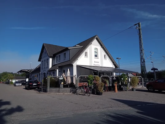 Gaststätte Gleis 3 in Augustfehn Das Bild zeigt die Gaststätte "Gleis 3" in Augustfehn. Das weiße Gebäude im traditionellen Design hebt sich mit einem markanten, dunklen Satteldach hervor. Davor sind Fahrräder und Pflanzen zu sehen, was eine gemütliche, einladende Atmosphäre schafft.