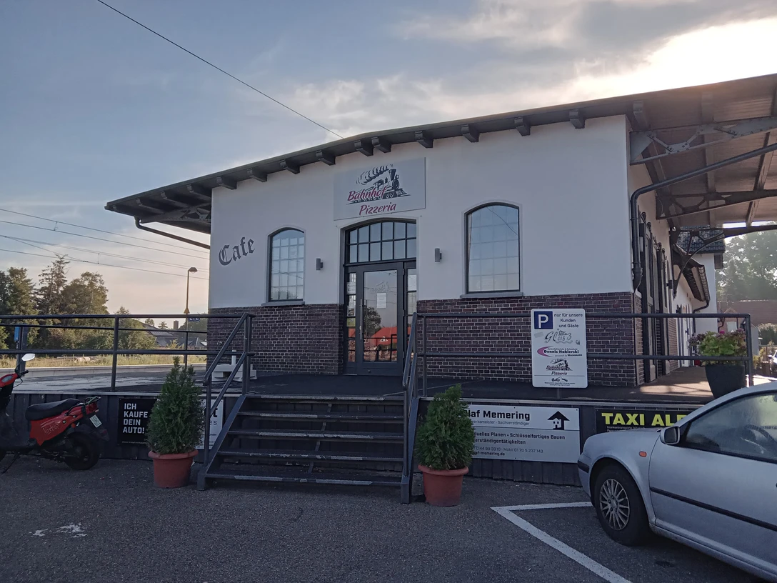 Bahnhof Pizzeria in Augustfehn Das Bild zeigt die Bahnhof Pizzeria in Augustfehn, ein modernes Gebäude mit großen Fenstern und rotem Backstein. Vor dem Eingang führen Stufen hinauf zur Tür, flankiert von dekorativen Blumenkübeln. Im Vordergrund sind Parkplätze und ein Schild mit Informationen sichtbar.