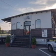 Bahnhof Pizzeria in Augustfehn Das Bild zeigt die Bahnhof Pizzeria in Augustfehn, ein modernes Gebäude mit großen Fenstern und rotem Backstein. Vor dem Eingang führen Stufen hinauf zur Tür, flankiert von dekorativen Blumenkübeln. Im Vordergrund sind Parkplätze und ein Schild mit Informationen sichtbar.
