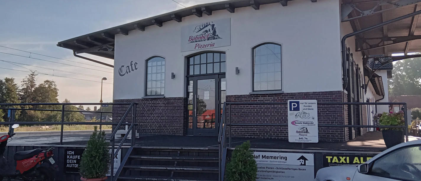 Bahnhof Pizzeria in Augustfehn Das Bild zeigt die Bahnhof Pizzeria in Augustfehn, ein modernes Gebäude mit großen Fenstern und rotem Backstein. Vor dem Eingang führen Stufen hinauf zur Tür, flankiert von dekorativen Blumenkübeln. Im Vordergrund sind Parkplätze und ein Schild mit Informationen sichtbar.