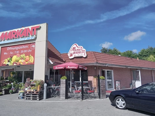 Bäckerei Musswessels in Apen Das Bild zeigt die charmante Fassade der Bäckerei Musswessels in Apen mit einem roten Ziegeldach und Außensitzplätzen.
