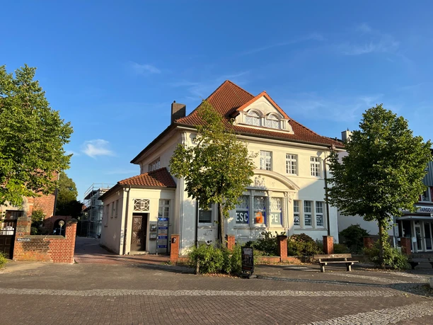 A large, white building with a red roof and escape room signs on the façade, surrounded by trees.