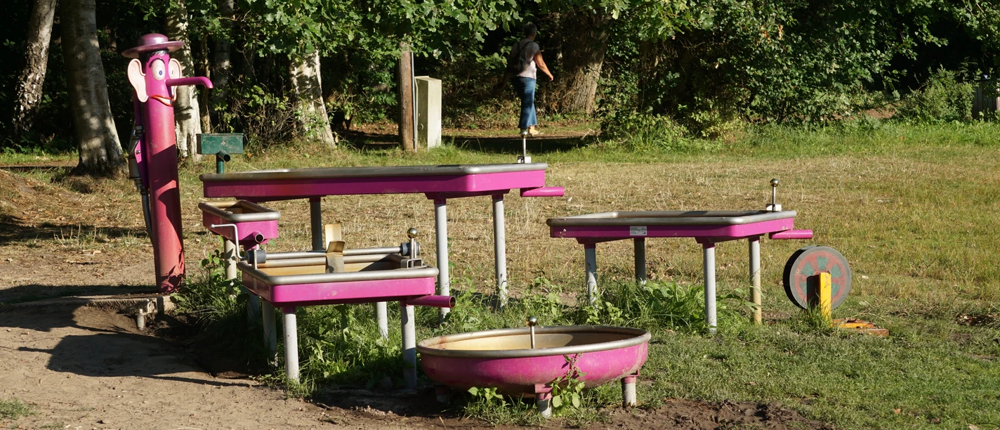 Wasserspielplatz am Lopausee Pinkfarbene Wasser-Matschanlage mit Pumpenfigur und Becken auf einer Wiese am Waldrand.