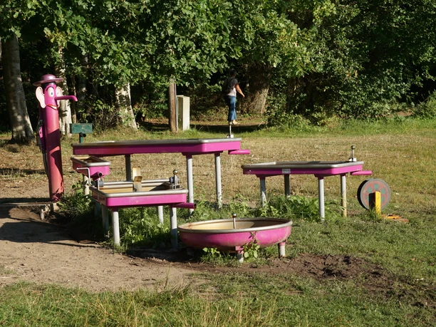 Wasserspielplatz am Lopausee Pinkfarbene Wasser-Matschanlage mit Pumpenfigur und Becken auf einer Wiese am Waldrand.
