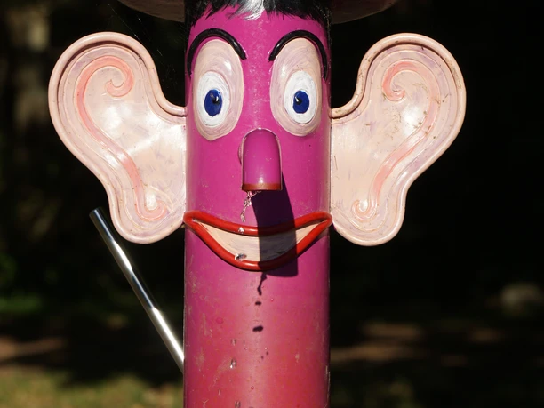 DSC05454.JPG Pinkfarbene Wasserpumpe in Form eines Gesichts mit großen Ohren, aus dessen Nase Wasser tropft.