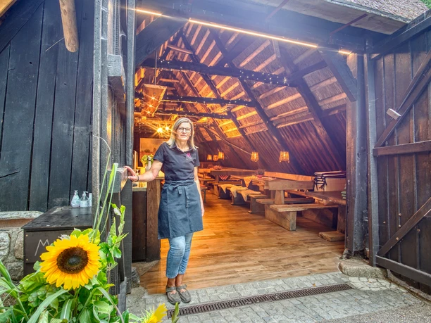 schafstall-amelinghausen-2023 Eine Frau steht im Eingang eines historischen, reetgedeckten Schafstalls mit Holztischen.
