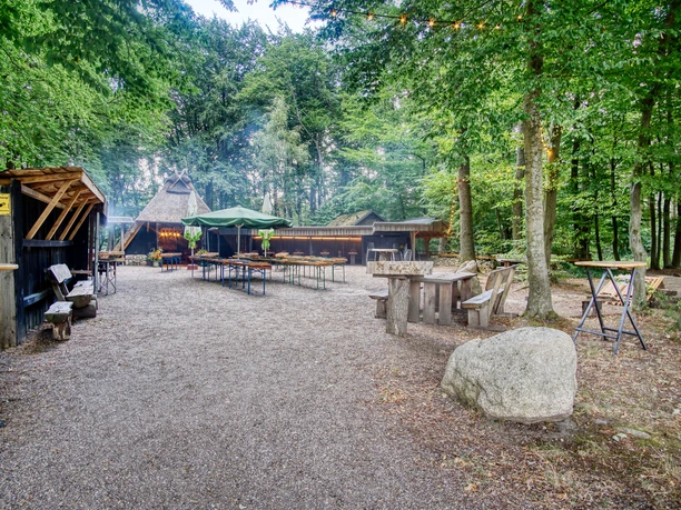 schafstall-hof-amelinghausen-2023 Außenbereich des Schafstalls Amelinghausen mit Tischen und Bänken unter Bäumen am Waldrand.