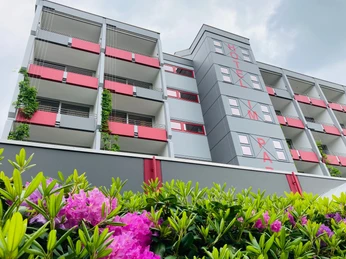 Moderne Hotel Fassade mit roten Balkonen und grüner Bepflanzung, umgeben von blühenden Sträuchern.