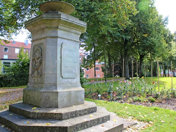 A stone memorial with a relief and an inscription, surrounded by trees and flower beds in the park.