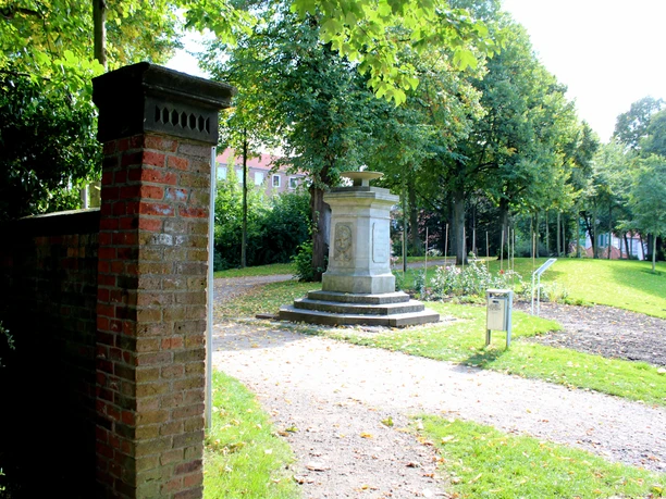 Gedenkstätte im Park mit steinerner Säule, umgeben von Bäumen und einem Weg, der durch die Anlage führt.