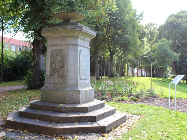 Ein Denkmal aus Stein, umgeben von grünem Rasen und Bäumen, mit einer Inschrift auf der Vorderseite.