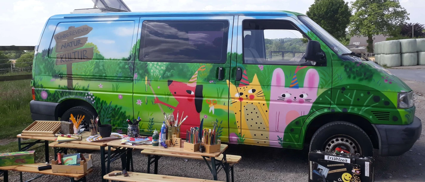 Erzähl- und Papierwerkstatt Mettmann Ein bunt bemalter Van in ländlicher Umgebung, flankiert von Holzbänken und Kunstmaterialien.