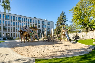 Drachenspielplatz in Ratingen Spielplatz mit Holzstrukturen, Rutsche und Sand, gelegen vor einem modernen Gebäude und umgeben von grünen Bäumen.