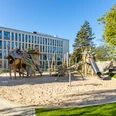 Drachenspielplatz in Ratingen Spielplatz mit Holzstrukturen, Rutsche und Sand, gelegen vor einem modernen Gebäude und umgeben von grünen Bäumen.