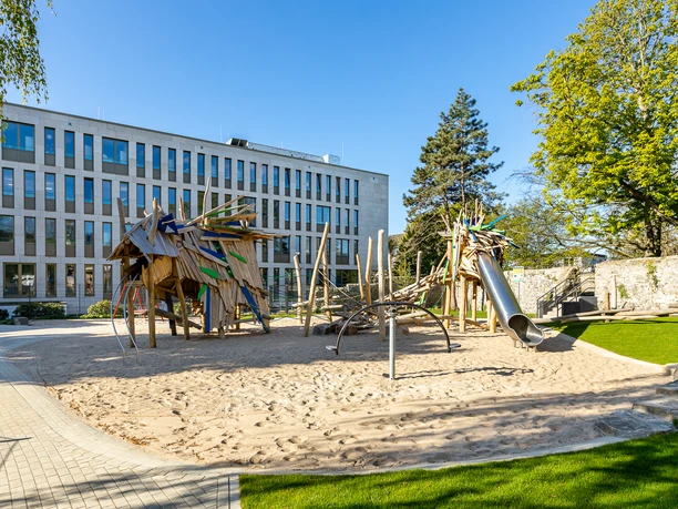 Drachenspielplatz in Ratingen Spielplatz mit Holzstrukturen, Rutsche und Sand, gelegen vor einem modernen Gebäude und umgeben von grünen Bäumen.