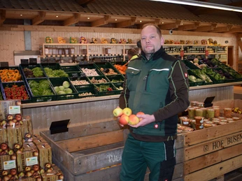 Bauerngarten Benninghoven Ein Landwirt präsentiert stolz frische Äpfel inmitten eines Bauernladens mit reichhaltigem Angebot.