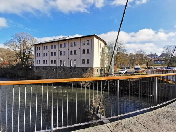 Neues Baumwolllager Ansicht eines historischen weißen Gebäudes an einem Fluss, mit moderner Brücke im Vordergrund.