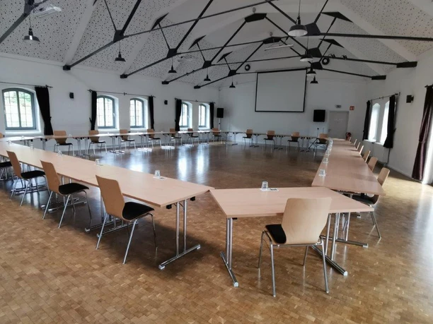 Innenansicht Heller Konferenzraum mit hölzernem Boden, U-förmiger Tischanordnung und moderner Beleuchtung.