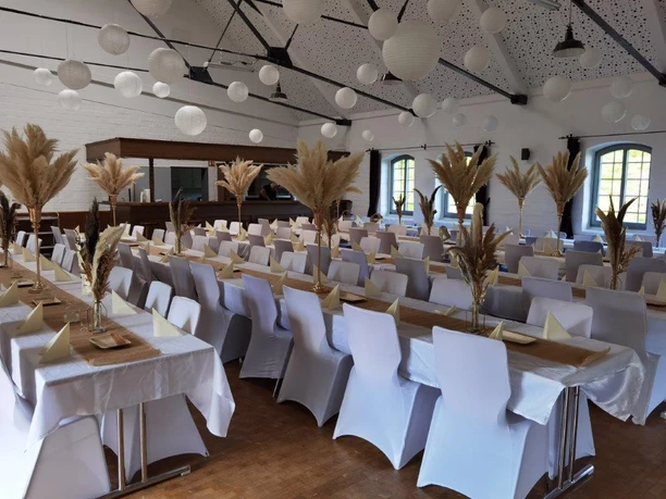 Innenansicht Großer Festsaal mit langen, weiß eingedeckten Tafeln und Pampasgras-Dekoration unter weißen Lampions.