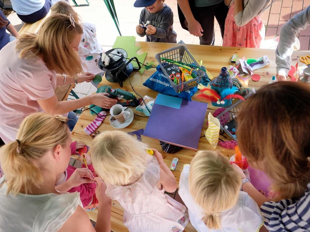Kinder basteln konzentriert an einem Tisch, umgeben von bunten Materialien und Erwachsenen.