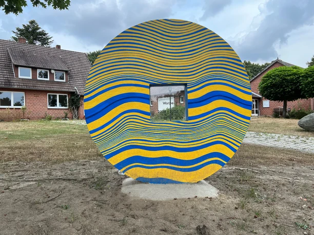 A sculpture in the shape of a circular element with blue and yellow wavy lines, placed on a patch of grass, with a small tree and brick buildings in the background.