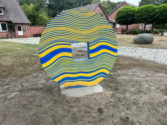 Skulptur aus bunten, wellenförmigen Streifen mit zentraler Widmung, umgeben von Backsteinhäusern.