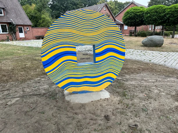 Skulptur Schlussstein Skulptur aus bunten, wellenförmigen Streifen mit zentraler Widmung, umgeben von Backsteinhäusern.