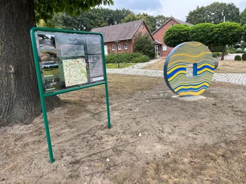 Runde Skulptur am Wegesrand inmitten einer grünen Parkanlage und einem Infotafel daneben.