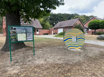 Skulptur Schlussstein Eine bunte, wellenförmige Skulptur neben einem Informationstafel auf einem ländlichen Platz.