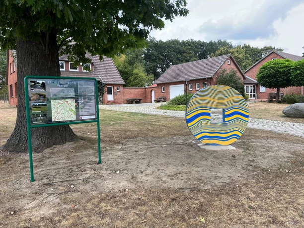 Skulptur Schlussstein Eine bunte, wellenförmige Skulptur neben einem Informationstafel auf einem ländlichen Platz.