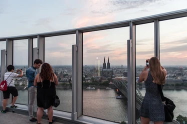 KölnTriangle Personen genießen den atemberaubenden Blick von der Aussichtsplattform des KölnTriangle über die Stadt Köln, die im Abendlicht erstrahlt. Der Panoramablick umfasst den majestätischen Kölner Dom und die glitzernde Rheinlandschaft, was ein unvergessliches Erlebnis bietet.