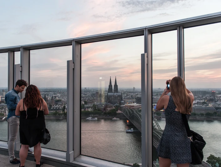 KölnTriangle Personen genießen den atemberaubenden Blick von der Aussichtsplattform des KölnTriangle über die Stadt Köln, die im Abendlicht erstrahlt. Der Panoramablick umfasst den majestätischen Kölner Dom und die glitzernde Rheinlandschaft, was ein unvergessliches Erlebnis bietet.Visitors enjoy the breathtaking view from the viewing platform of the KölnTriangle over the city of Cologne, which shines in the evening light. The panoramic view includes the majestic Cologne Cathedral and the glittering Rhine landscape, offering an unforgettable experience.