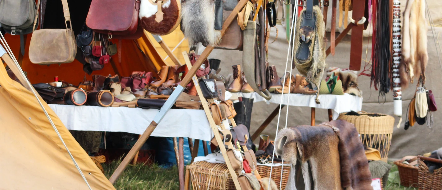 Marktstand mit mittelalterlichen Waren wie Ledertaschen, Fellkleidung, Holzgeschirr und Körben.