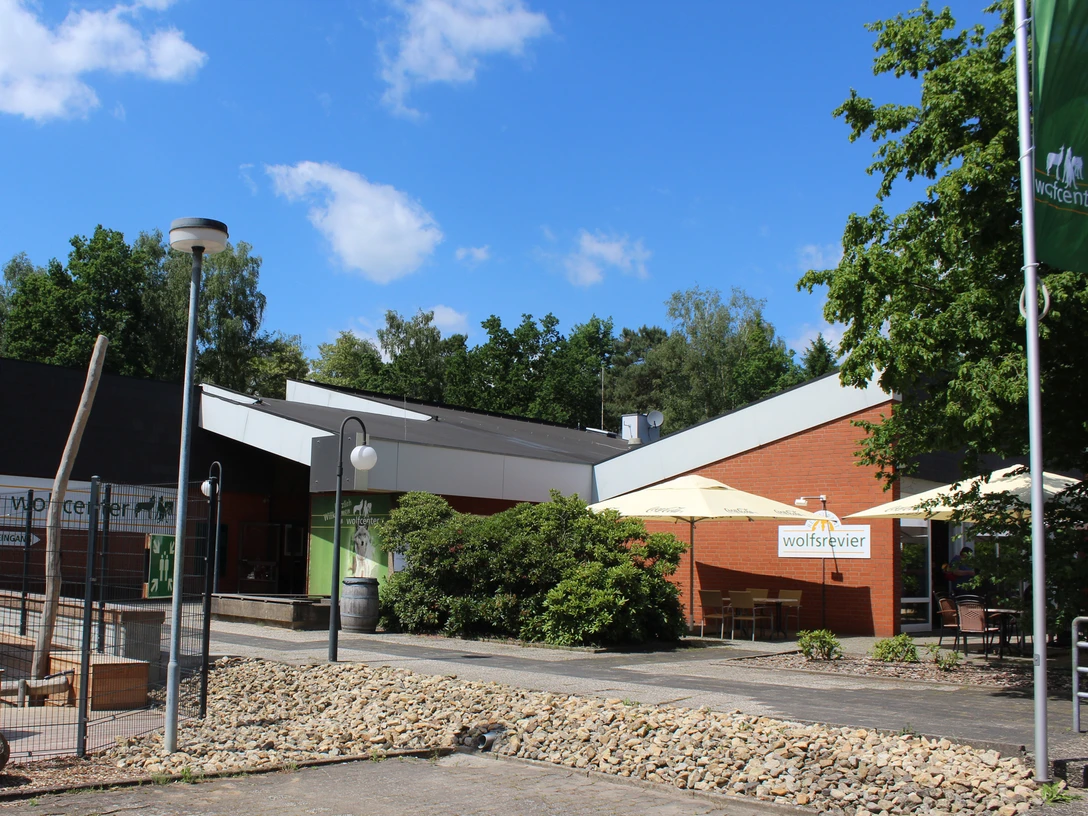 Moderner Eingangsbereich des Wolfcenters mit begrünten Flächen, überdachten Sitzmöglichkeiten und blauen Himmel.Modern entrance area of the Wolfcenter with green areas, covered seating and blue sky.Moderne indgangsparti til Wolfcenter med grønne områder, overdækkede siddepladser og blå himmel.Modern entreegebied van het Wolfcenter met groene zones, overdekte zitplaatsen en blauwe lucht.