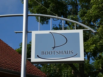 Ein Schild mit einem großen stilisierten "B" und dem Wort "Bootshaus" vor einem klaren Himmel.