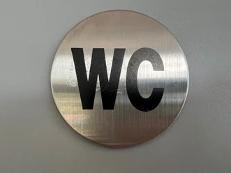 Silbernes, rundes WC-Schild mit schwarzer Aufschrift vor grauem Hintergrund, schlichtes Design.