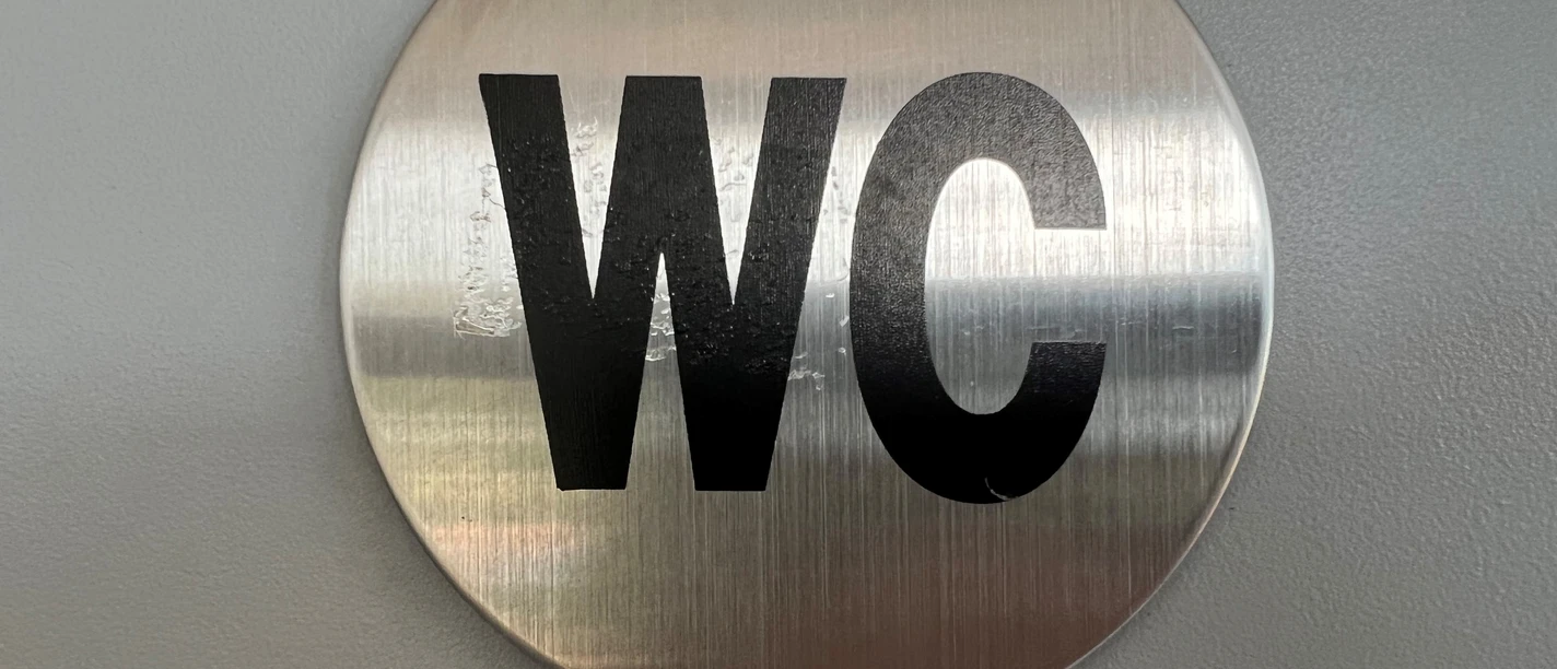 Silbernes, rundes WC-Schild mit schwarzer Aufschrift vor grauem Hintergrund, schlichtes Design.