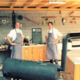 Außenküche in der Kleinen Schweiz, Velbert Zwei Personen in Schürzen stehen in einer rustikalen Außenküche mit Grill und Dekorationen.