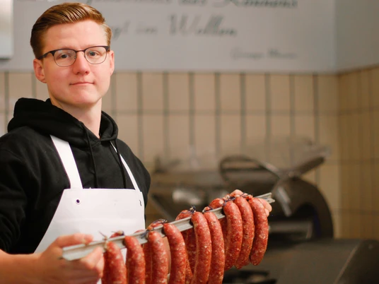 Eigene Wurstherstellung in der Metzgerei Kluke, Mettmann Metzger mit Schürze hält eine Stange frischer Bratwürste in einer traditionellen Metzgerei.