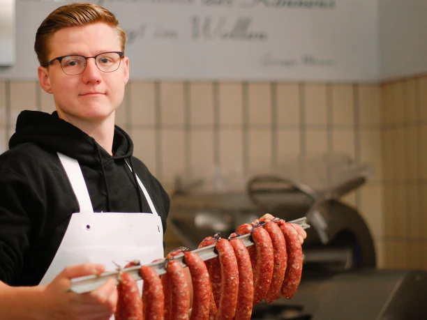 Eigene Wurstherstellung in der Metzgerei Kluke, Mettmann Metzger mit Schürze hält eine Stange frischer Bratwürste in einer traditionellen Metzgerei.